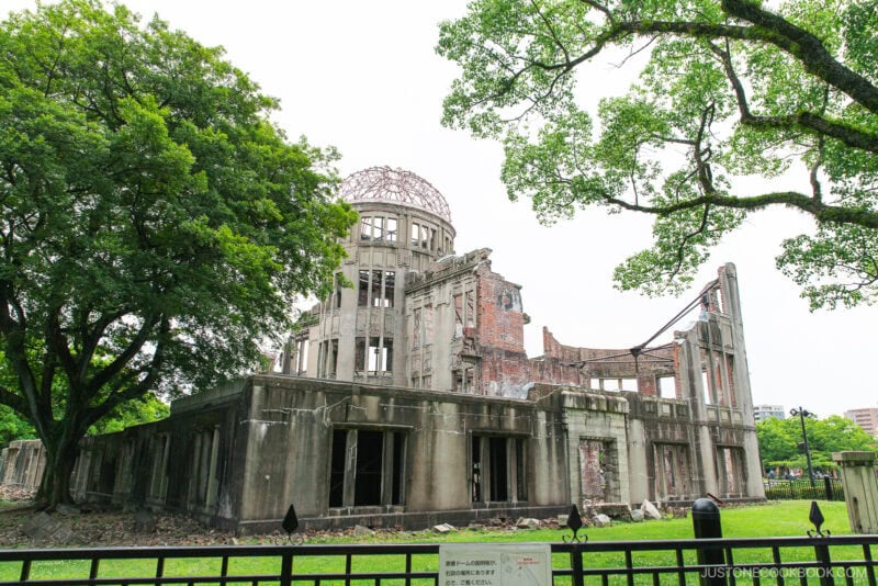 The Hiroshima Peace Memorial, a partially destroyed concrete building with a domed framework, stands surrounded by green trees and grass within Hiroshima Peace Memorial Park under a bright, cloudy sky.