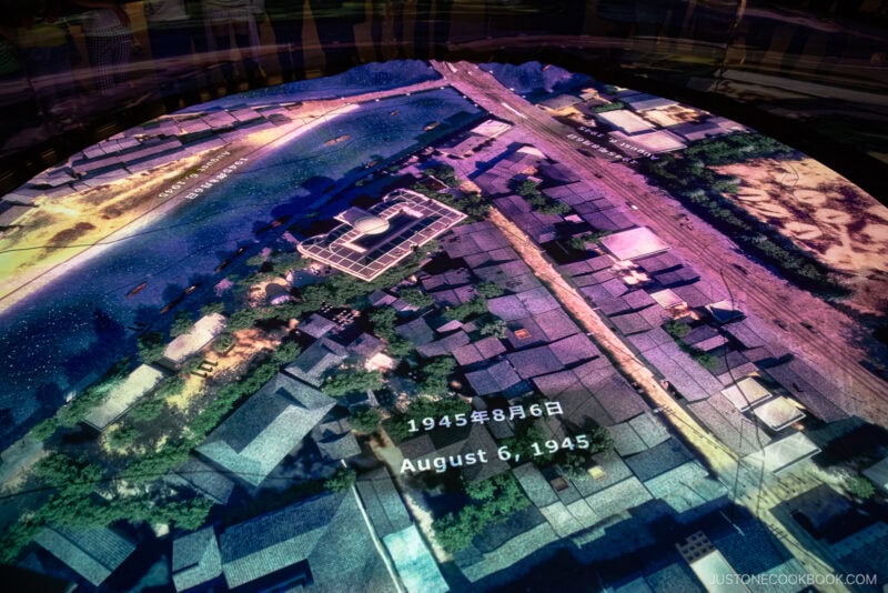 A digital display in Hiroshima Peace Memorial Park shows an aerial map of Hiroshima on August 6, 1945, with the date in both Japanese and English, depicting buildings and streets before the atomic bombing.