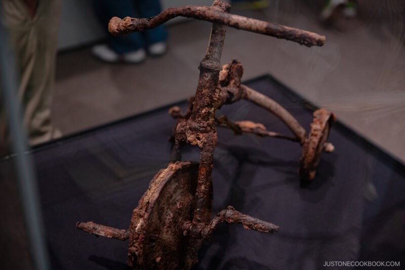 A rusted, damaged tricycle is displayed in a glass case on a black platform at Hiroshima Peace Memorial Park. The tricycle’s corroded frame and wheels stand out against the blurred figures and shoes in the background.