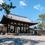 Horyu-ji (法隆寺) – The Oldest Wooden Buildings in the World Nara Horyu-ji’s traditional Japanese temple gate features a tiled roof, wooden pillars, and stone steps, nestled among pine trees beneath a bright blue sky with scattered clouds.