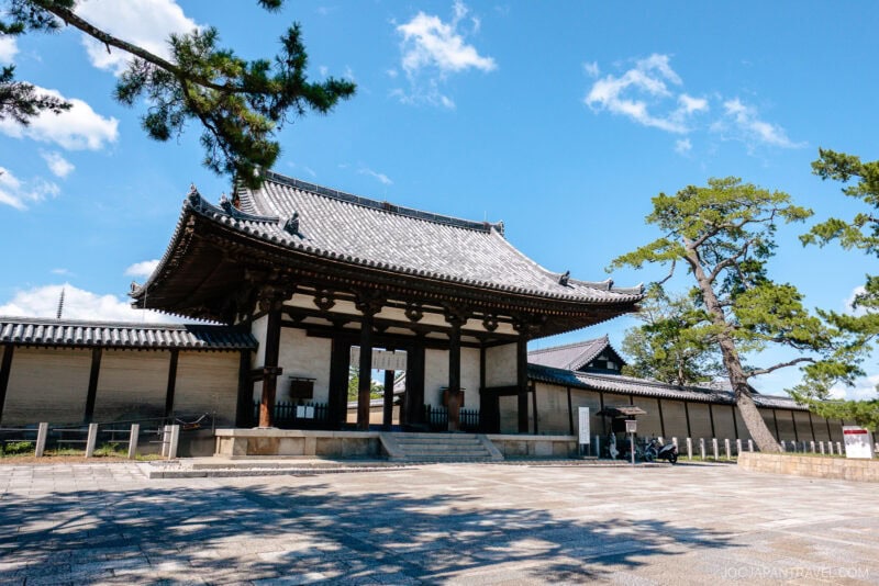 Nara Horyu-ji’s traditional Japanese temple gate features a tiled roof, wooden pillars, and stone steps, nestled among pine trees beneath a bright blue sky with scattered clouds.