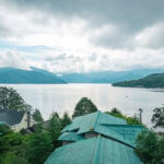 A peaceful lakeside scene with green-roofed buildings, including Hoshino Resorts KAI Nikko in the foreground, surrounded by lush trees and distant mountains under a partly cloudy sky reflected in the calm lake.