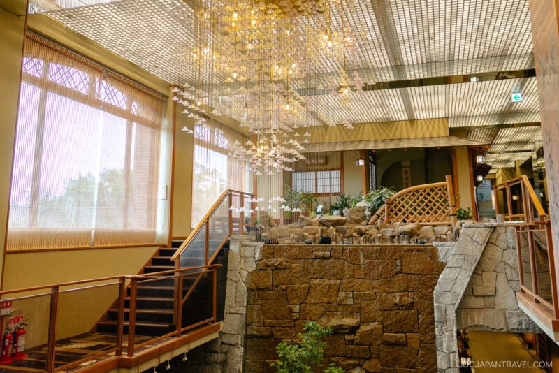 A spacious, sunlit interior at Hotel Kaneyamaen Yamanashi features large windows, a grand chandelier, wooden railings, stone stairs, and an indoor rock garden, creating a serene and elegant atmosphere.