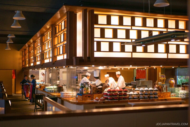 A modern Japanese restaurant interior at Hotel Kaneyamaen Yamanashi features chefs in white uniforms preparing food behind a counter, stacks of bowls, and warm wooden decor; staff in patterned uniforms stand nearby.