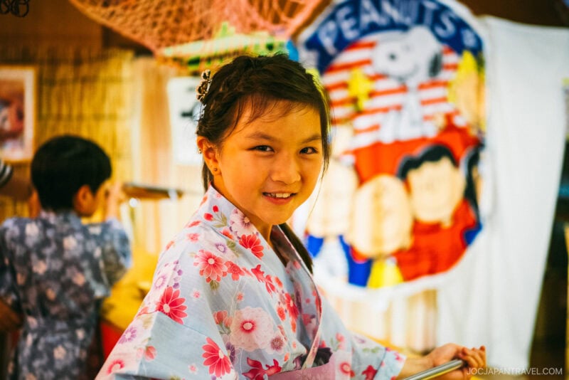 A young girl in a pink floral yukata smiles at the camera at Hotel Kaneyamaen Yamanashi; a Peanuts cartoon banner is in the background, with another child in a yukata standing nearby.