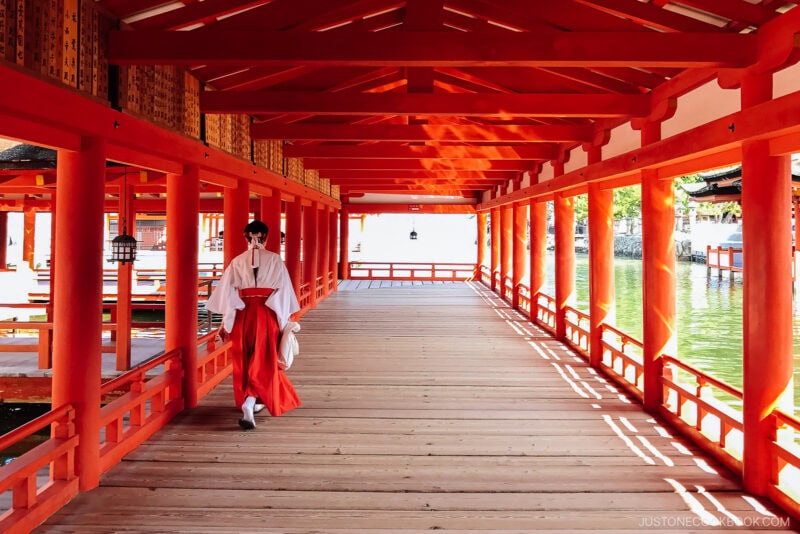 A person in traditional Japanese attire walks through a bright, red-painted wooden corridor at Itsukushima Shrine, sunlight streaming through the beams and reflecting on the polished floor of this iconic Shinto site.