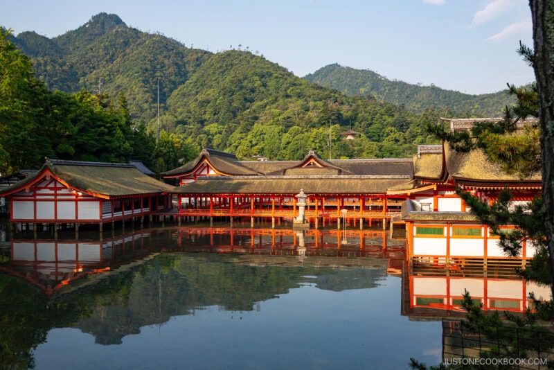 Traditional Japanese shrine buildings with red and white walls and curved roofs stand over calm water, reflecting the structures. Itsukushima Shrine rests peacefully as lush green mountains rise in the background under a clear sky.