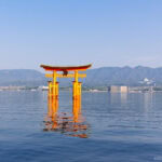 Itsukushima Shrine (厳島神社) – Miyajima’s Floating Icon of Japan A large, bright orange torii gate of Itsukushima Shrine stands in the water with distant mountains, city buildings, and a clear blue sky in the background. The gate’s reflection shimmers on the calm surface of the water.