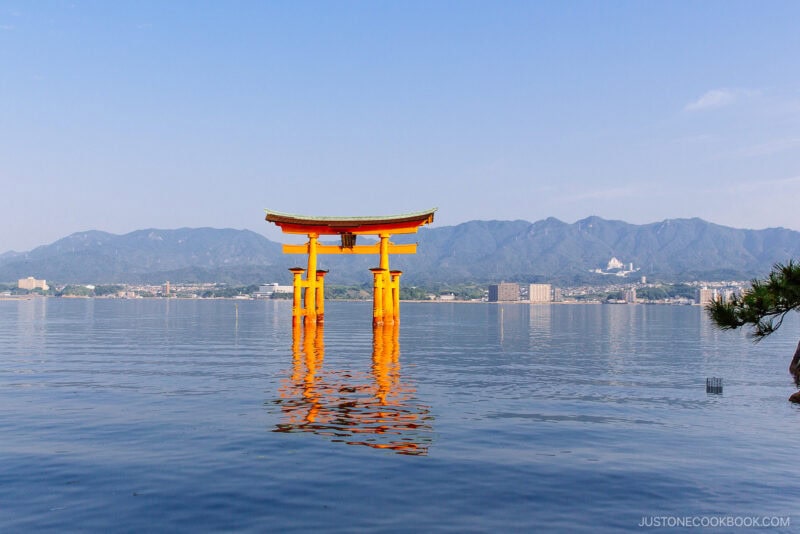 A large, bright orange torii gate of Itsukushima Shrine stands in the water with distant mountains, city buildings, and a clear blue sky in the background. The gate&rsquo;s reflection shimmers on the calm surface of the water.