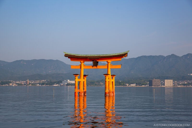A large, traditional red torii gate of Itsukushima Shrine stands in the water, with mountains and buildings visible in the background under a clear blue sky.