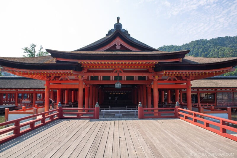 A view of the entrance to the famed Itsukushima Shrine, showcasing red wooden beams, intricate roof details, and a wooden walkway, all set against a backdrop of lush trees and blue sky.