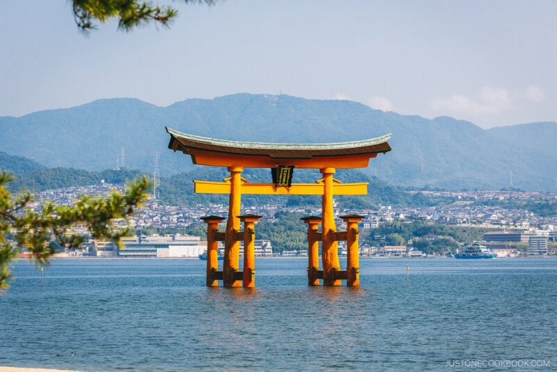 The iconic red torii gate of Itsukushima Shrine stands in the water, framed by mountains and a coastal town on Miyajima Island, Japan, beneath a clear sky.