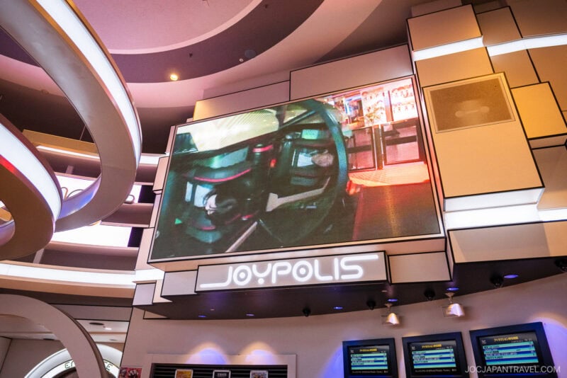 A large screen displays a video above the entrance to Joypolis DECKS, a futuristic indoor amusement park featuring curved white architectural elements and vibrant electronic displays.