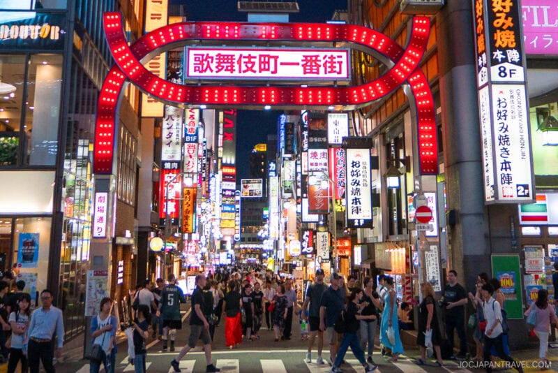 A busy street in Tokyo’s Kabukicho district at night, crowded with people and illuminated by vibrant neon signs in Japanese. The large red Kabukicho Ichibangai archway sign is prominent above the bustling entrance.