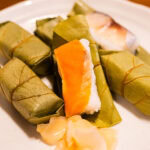 Kakinoha-zushi (柿の葉寿司) – Nara’s Leaf-Wrapped Sushi Tradition A plate with Hiraso Kakinoha-zushi—wrapped persimmon leaf sushi, some pieces unwrapped to reveal rice and fish—served alongside a small portion of pickled ginger.