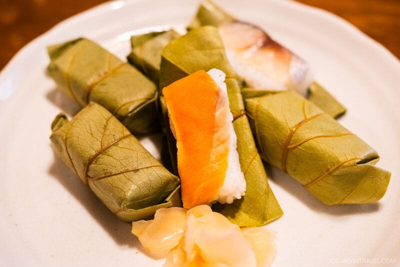 A plate with Hiraso Kakinoha-zushi—wrapped persimmon leaf sushi, some pieces unwrapped to reveal rice and fish—served alongside a small portion of pickled ginger.