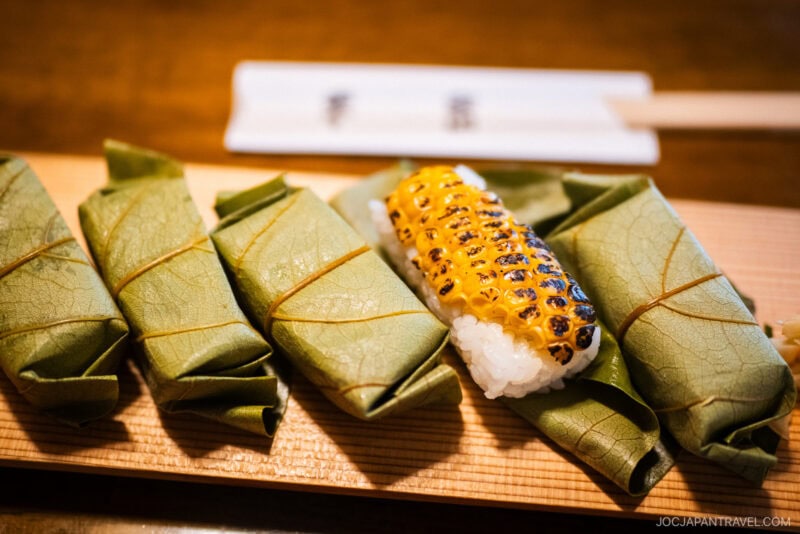 Four pieces of Hiraso Kakinoha-zushi, wrapped in green leaves, sit on a wooden board beside sushi topped with grilled corn. Chopsticks and a holder are softly blurred in the background.