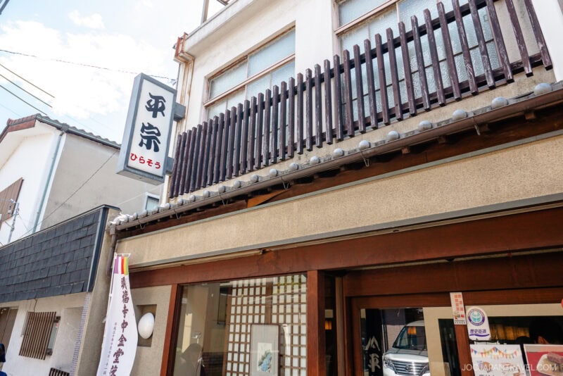 The exterior of a Japanese building with a wooden balcony and vertical slats, featuring a white Hiraso Kakinoha-zushi sign with black Japanese characters and smaller red text, under a clear sky.