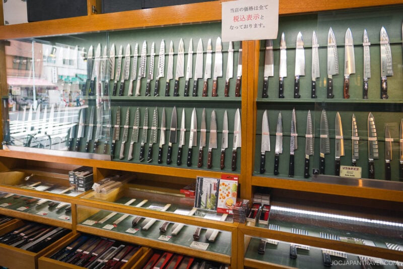 A glass display case in Kappabashi Kamata Knife Shop showcases a wide variety of Japanese kitchen knives, neatly arranged in rows, with boxed knives and sharpeners on the lower shelves and a sign in Japanese above.