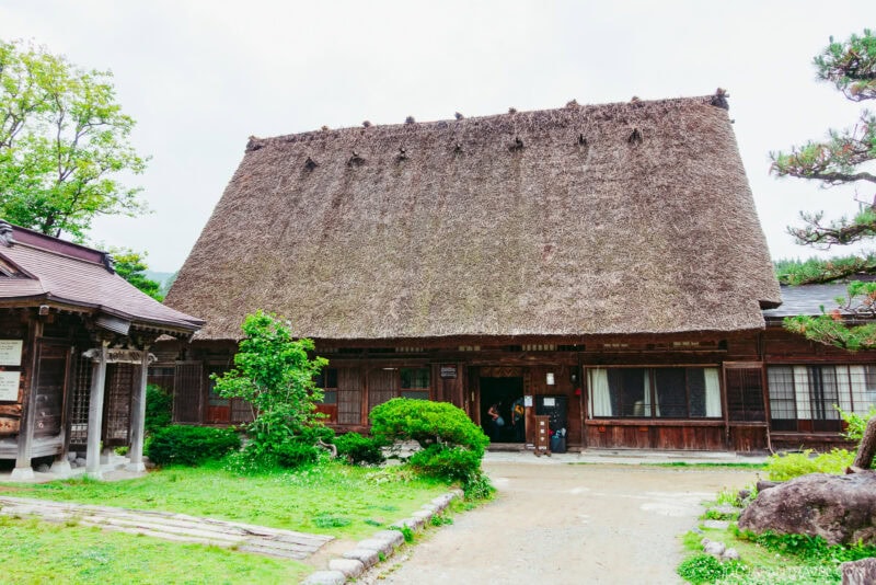 A traditional Japanese house, like Kanda House Shirakawa-go, with a steep thatched roof, wooden walls, and a garden filled with green shrubs and trees in front, rests under a cloudy sky.