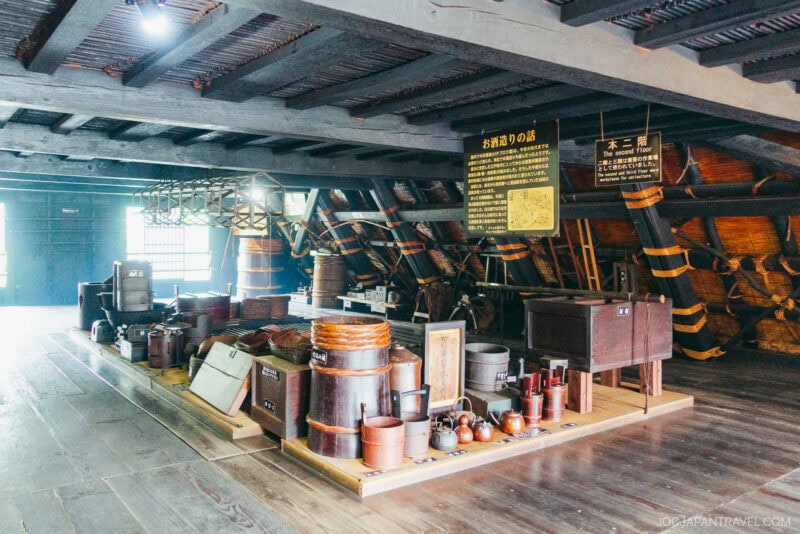 A museum exhibit at Kanda House Shirakawa-go displays traditional Japanese wooden tools, pots, and household items on a polished wooden floor, with informational signs and dim lighting inside a historic building.