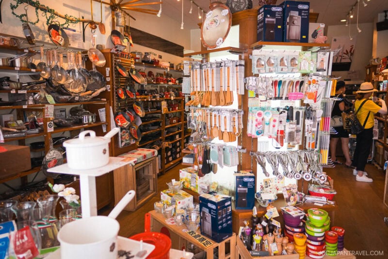 A brightly lit shop, Kappabashi Dr. Goods, filled with kitchenware, utensils, pots, pans, and various cooking tools displayed on shelves and racks. Two shoppers are browsing in the background.
