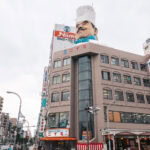A multi-story building in Tokyo’s Kappabashi district features a giant chef’s head wearing a white hat on its roof. The facade displays the Niimi sign and is surrounded by other urban buildings under a cloudy sky.