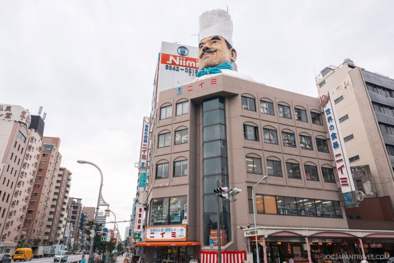 A multi-story building in Tokyo&rsquo;s Kappabashi district features a giant chef&rsquo;s head wearing a white hat on its roof. The facade displays the Niimi sign and is surrounded by other urban buildings under a cloudy sky.