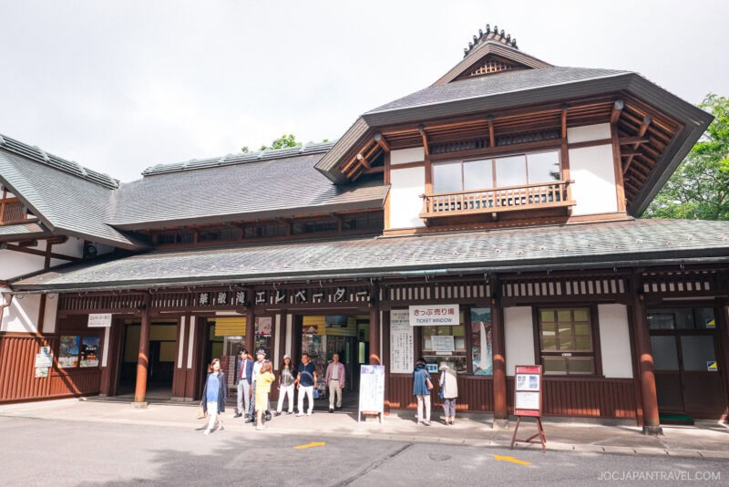 A group of people stand outside a traditional Japanese building with a peaked tiled roof and wooden details; signs above the entrance indicate it is a train station, a common gateway for visitors heading to Nikko Kegon Falls.