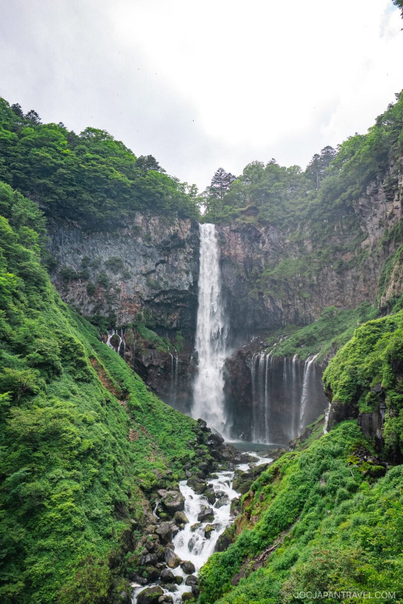 Nikko Kegon Falls cascades down a rocky cliff, surrounded by lush green trees and vegetation, with white mist and water flowing over rocks at the base under a bright, cloudy sky.