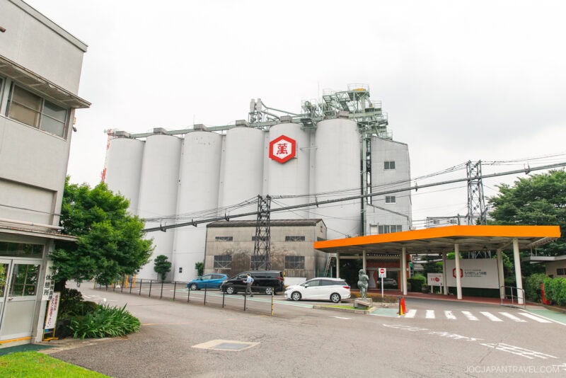 A large industrial building with multiple white silos, a red hexagonal logo, parked cars, and surrounding trees under an overcast sky—the site of the Kikkoman Factory Tour.