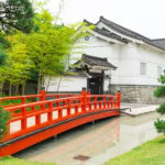 A vibrant red arched bridge crosses over a narrow stream in front of a traditional white Japanese building, surrounded by lush green trees and bamboo—reminiscent of the serene scenery found on a Kikkoman Factory Tour.