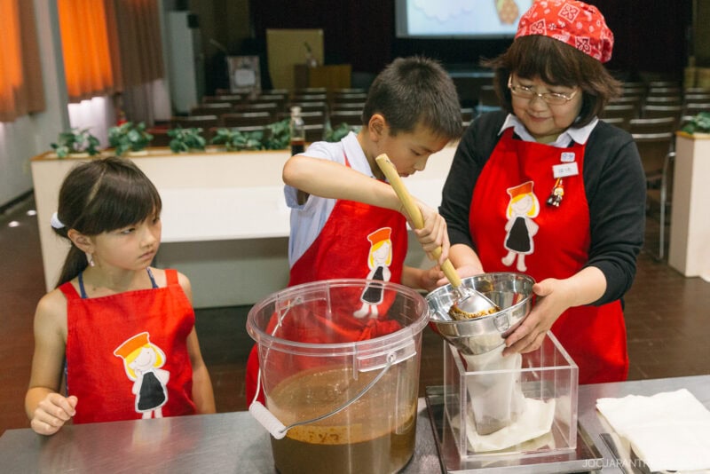 A woman and two children in red aprons work together to pour a brown liquid mixture through a cloth filter, echoing the careful tradition seen on a Kikkoman Factory Tour, as the girl watches the boy stir and the woman assists.