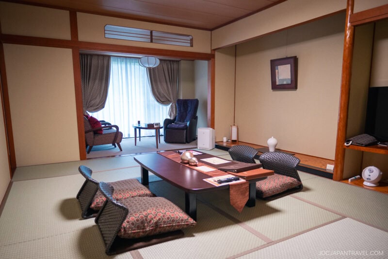 A traditional Japanese tatami room at Kinugawa Onsen features a low table, floor cushions, and a tea set. Two armchairs by the window with curtains complement the wooden accents and minimalistic decor.