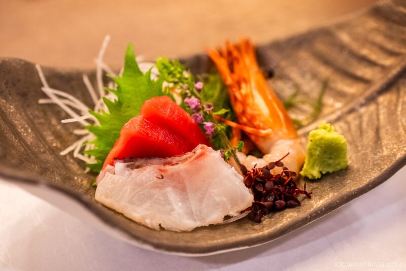 A plate of sashimi at Kinugawa Onsen features slices of white fish, tuna, shrimp, garnished with shiso leaf, shredded daikon, tiny purple flowers, and a dollop of wasabi on a textured ceramic dish.