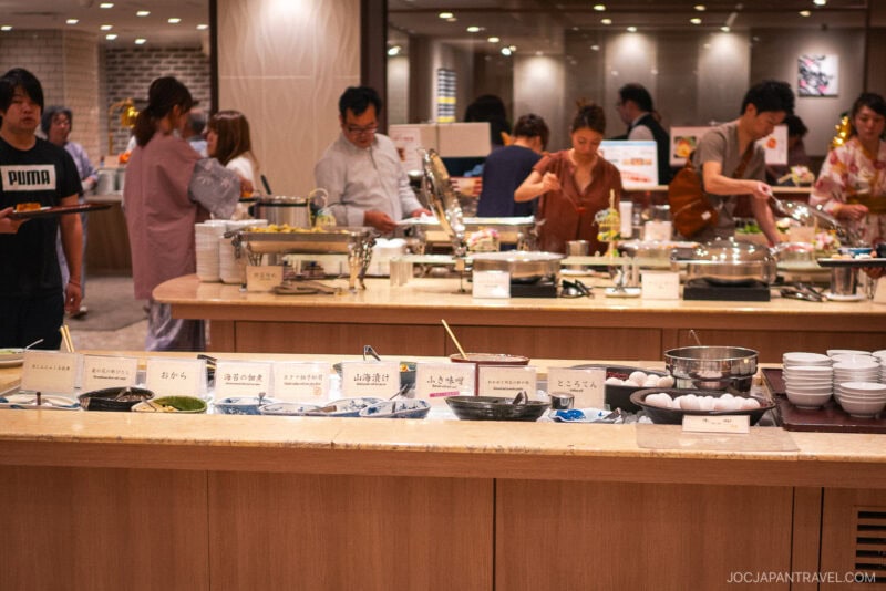 A buffet restaurant in Kinugawa Onsen with people serving themselves food. Various dishes, bowls, and trays are displayed on the counter with Japanese labels. The atmosphere is busy and bright, with several diners in the background.