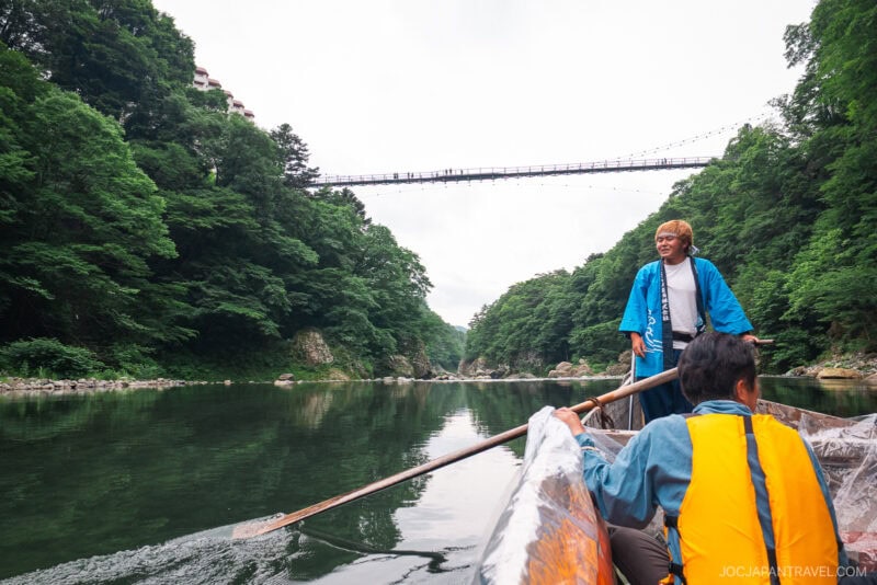 Two people ride a wooden boat along the calm Kinugawa Onsen river, surrounded by lush green forest. One rows while the other sits in a yellow life jacket. A tall suspension bridge spans overhead.
