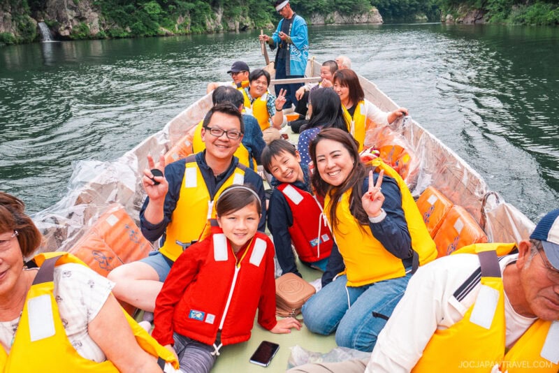 A group of smiling people wearing yellow life jackets sit on a boat at Kinugawa Onsen, posing for a photo with peace signs. The boat drifts on a calm river surrounded by lush greenery and rocky cliffs.
