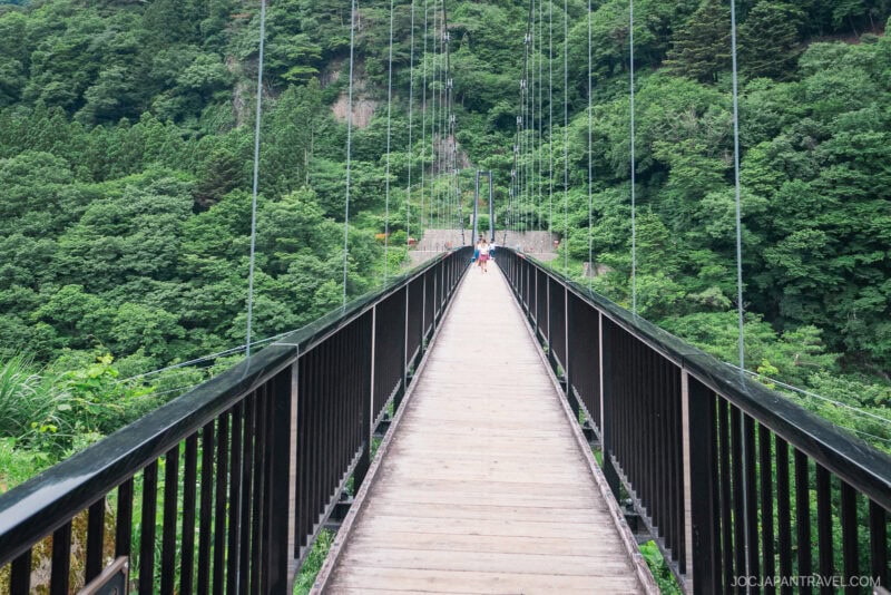 A long suspension bridge with black railings stretches into a lush, green forest near Kinugawa Onsen. Two people are walking in the distance, surrounded by tall trees and dense foliage.