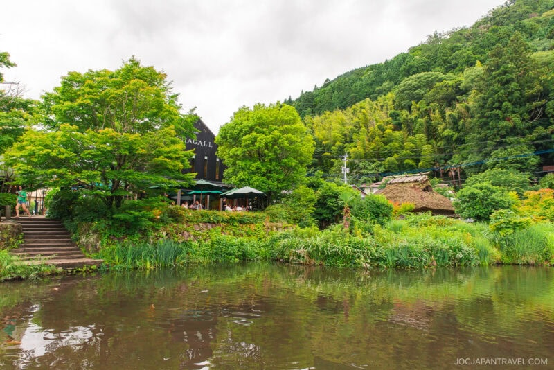 A scenic view of Kirin Lake, surrounded by lush greenery, with a caf&eacute; and outdoor seating on the left and a thatched-roof house in the background, set against forested hills under a cloudy sky.