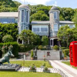 A stone mansion with two round towers, reminiscent of Kitano Ijinkan, stands among lush greenery. In the garden, a boar statue and classic red British phone booth sit amid flowers leading to the ornate house under a bright blue sky.
