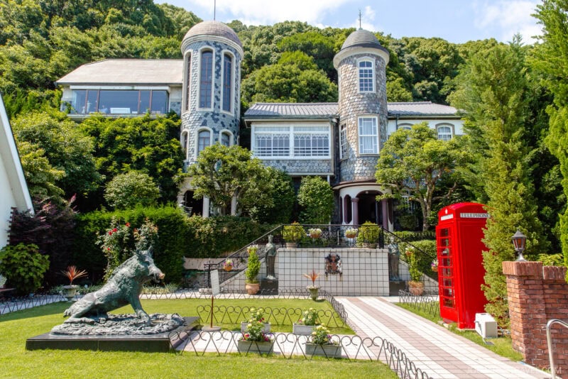 A stone mansion with two round towers, reminiscent of Kitano Ijinkan, stands among lush greenery. In the garden, a boar statue and classic red British phone booth sit amid flowers leading to the ornate house under a bright blue sky.