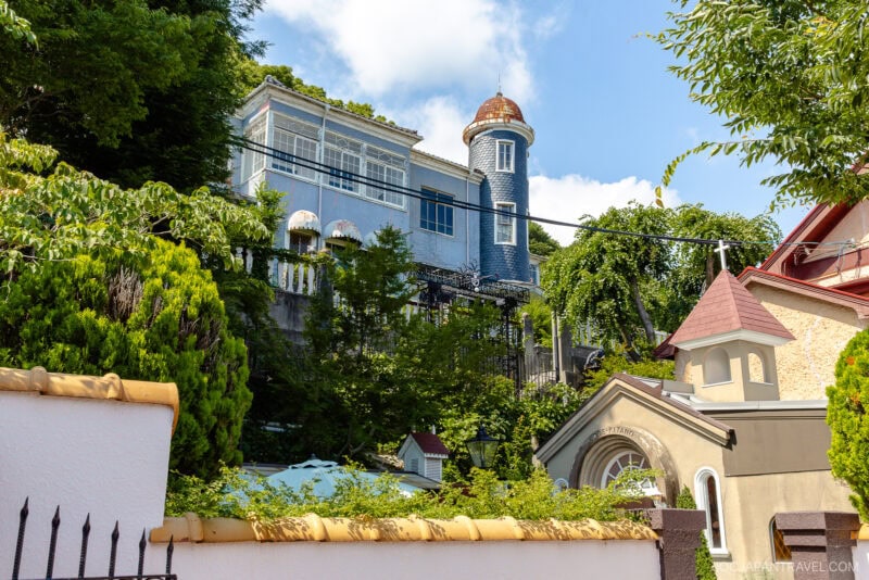A whimsical blue building with a round turret, reminiscent of Kitano Ijinkan, stands on a hillside surrounded by greenery, next to a cream-colored house with a pointed roof under a partly cloudy sky.