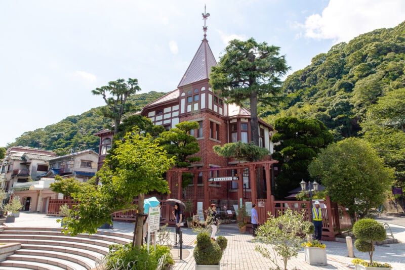 A red-brick Western-style building with a pointed roof stands amid lush greenery and trees in Kitano Ijinkan, with people walking and sitting nearby on a sunny day. Green hills rise in the background under a blue sky.
