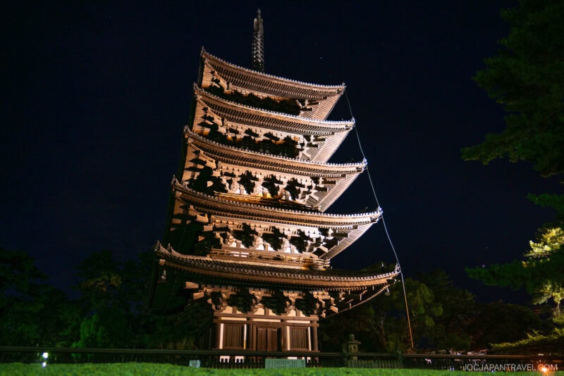 A tall, illuminated five-story wooden pagoda at Nara Kofukuji stands against a dark night sky, surrounded by trees. The structure is intricately designed and lit with warm lights, highlighting its traditional Japanese architecture.