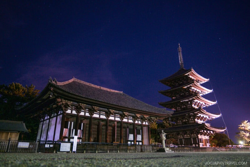 A traditional Japanese temple and the five-story Nara Kofukuji pagoda glow under a clear, starry sky. Surrounded by trees and enclosed by a low fence, these iconic structures create a serene nighttime scene.