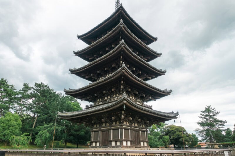 A traditional five-story wooden pagoda at Nara Kofukuji stands amid green trees under a cloudy sky, showcasing intricate architecture and elevated eaves.