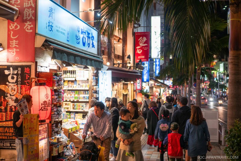 A busy street in Japan at night, lined with glowing shop signs and stores. Shoppers and families stroll along Kokusai Dori under palm trees, creating a lively and vibrant atmosphere.