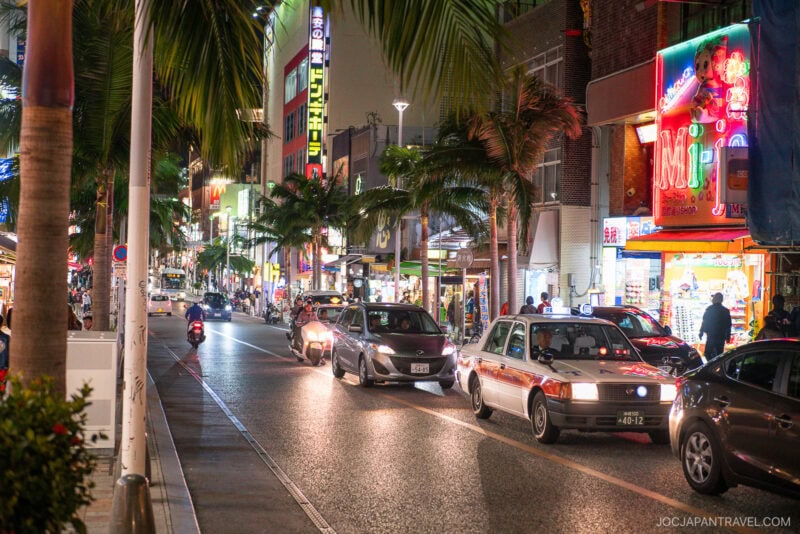 A lively city street at night in Japan, lined with palm trees and neon signs. Shops and restaurants along Kokusai Dori bustle as cars drive by and people stroll the vibrant sidewalks.