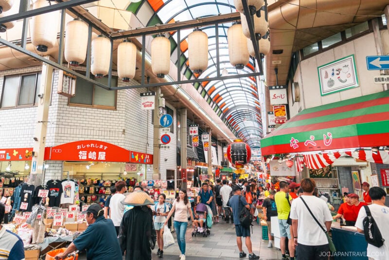 A busy covered market street in Japan, Kuromon Ichiba Market features hanging lanterns, colorful signs, shops, and crowds of people shopping and walking. The market has a vibrant, lively atmosphere.